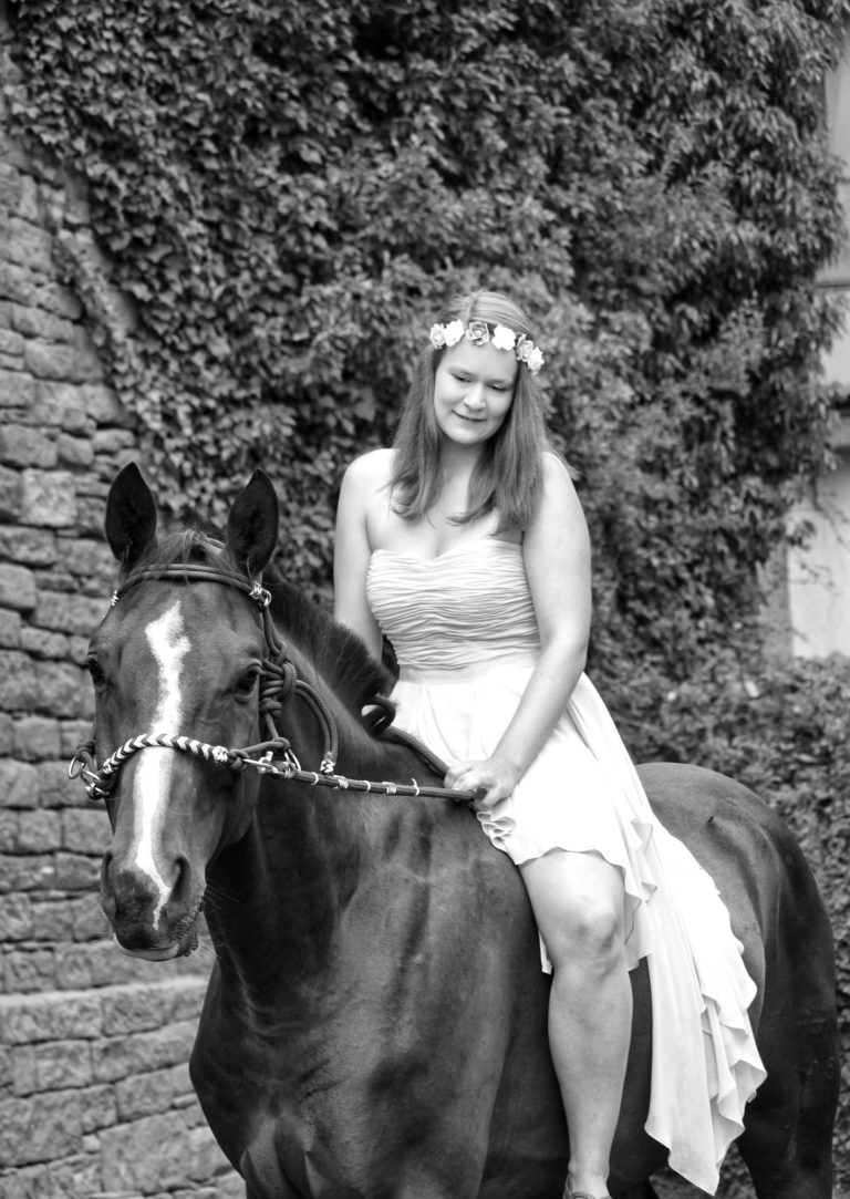 Inhaberin, wie sie auf ihrem ersten Pferd ohne Sattel sitzt. mit einem gebisslosen Zaum.
Sie hat ein Kleid an und einen Blumenkranz im Haar